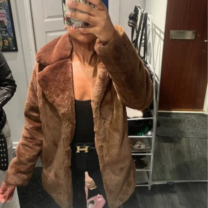 Brun pälsjacka med krage - Säljer en brun fusk pälsjacka med bred krage och rak passform. Jackan är fluffig och har en snygg oversized look, perfekt för att hålla sig varm och stilsäker. Färgen är mellanbrun och materialet känns mjukt och lyxigt.
