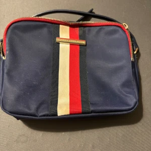 Blå axelväska Tommy Hilfiger - Snygg marinblå axelväska från Tommy Hilfiger med klassiska ränder i rött, beige och svart framtill. Väskan har guldfärgade dragkedjor och en stilren, boxig form. Perfekt för att bära det viktigaste med stil.