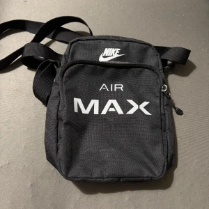 Nike Air Max svart axelväska - Snygg svart axelväska från Nike Air Max med vit logga och text framtill. Väskan har justerbar rem, dragkedja och är perfekt för att bära dina viktigaste grejer. Tillverkad i slitstark polyester och har en kompakt, smidig form.
