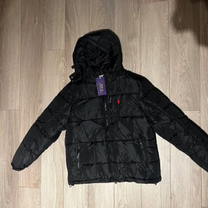 Svart pufferjacka från Polo Ralph Lauren - Svart pufferjacka från Polo Ralph Lauren med huva och dragkedja framtill. Jackan har två sidofickor med dragkedja och den klassiska röda Polo-loggan på bröstet. Perfekt för kalla dagar och riktigt snygg till streetwear-stilen.Kvitto finns 
