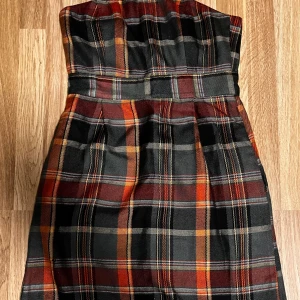 Rutig klänning b.young - Axelbandslös kortklänning från b.young i rutigt mönster med svarta, röda och orange toner. Klänningen har markerad midja och dragkedja där bak. Materialet är kraftigt och känns som bomullsmix med satinfoder. Perfekt för dig som vill sticka ut med en cool retrostil.