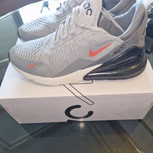 Nike Air 270 sneakers ljusgrå - Nike Air 270 sneakers i ljusgrå mesh med orange swoosh-detaljer. Skorna har en stor synlig Air-enhet i hälen, snörning och ventilerande ovandel. Snygg och modern design med rund tå och platt sula, perfekt för dig som gillar sportig stil.