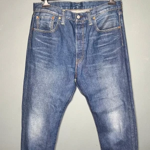 Levi's 501 CT blå jeans W34 L32 - Klassiska Levi's 501 CT jeans i blå denim med raka ben och femficksmodell. Snyggt slitna detaljer framtill och bak, ikonisk läderpatch på bakfickan och orange sömmar. Perfekt för dig som gillar tidlös streetstil.