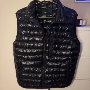 Svart dunväst Peak Performance M - Svart, glansig dunväst från Peak Performance i storlek M. Västen har quiltad design, hög krage och tre dragkedjeförsedda fickor framtill. Perfekt för lager-på-lager och snygg till både hoodie och t-shirt.