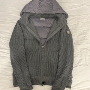 Grå stickad cardigan från Moncler - Snygg grå cardigan från Moncler med stickade ärmar och ribbade muddar. Jackan har quiltad baksida och huva i glansigt material, samt dragkedja framtill. Ikonisk Moncler-logga på ärmen. Perfekt för kyliga dagar när du vill vara både varm och stilren. Aldrig använd