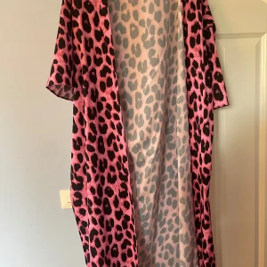 Långklänning med rosa leopardmönster - Statement långklänning i rosa med svart leopardmönster. Klänningen har korta ärmar och är tillverkad i ett lätt, flowy material som ger en cool och avslappnad vibe. Perfekt för dig som vill sticka ut och älskar djurmönster.