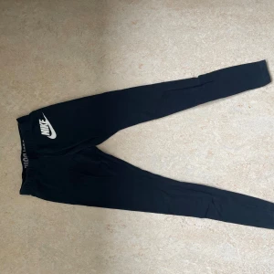 Svarta Nike mjukisbyxor med logga - Svarta mjukisbyxor från Nike med vit logga på vänster lår och texten 'JUST DO IT.' på insidan av midjan. Byxorna har elastisk midja och smal passform, perfekta för chill eller träning. Tillverkade i mjukt bomullsmaterial för extra komfort.