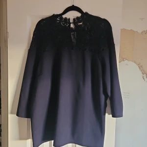 Mörkblå topp med spets från Holly & Whyte - Snygg mörkblå topp från Holly & Whyte med lång ärm och vacker spets upptill. Toppen har en loose passform och är tillverkad i mjuk polyesterblandning. Perfekt för dig som gillar stilrena detaljer och en feminin touch.