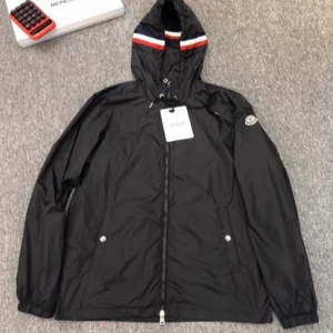 Moncler vindjacka - Helt ny och oanvänd svart vindjacka från Moncler med huva som har röd, vit och blå randdetalj. Jackan har dragkedja framtill, två fickor med knappar och Moncler-logga på ärmen. Perfekt för dig som gillar stilren och sportig look.