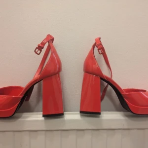 Röda chunky klackar från Bershka  - Säljer ett par röda chunky klackar från Bershka i storlek 38. Skorna har bred klack, rund tå och platåsula. De är gjorda i glansigt syntetmaterial och har justerbar rem runt vristen. Perfekta för dig som vill sticka ut med färg och cool retrostil  .