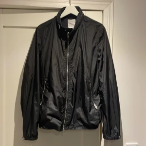  bomberjacka J.Lindeberg - Svart bomberjacka från J.Lindeberg i blankt syntetmaterial. Jackan har klassisk passform, ståkrage och silverfärgad dragkedja framtill samt vid fickorna. Perfekt för dig som gillar stilren och enkel design.