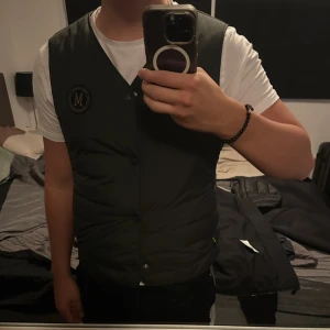 olive grön dunväst från Morris Stockholm(äkta) - Stilren svart dunväst från Morris Stockholm med rundad halsringning och tryckknappar framtill. Västen har ett broderat märke med guldigt M på bröstet och quiltad design. Perfekt för lager-på-lager och snygg till både hoodie och skjorta.