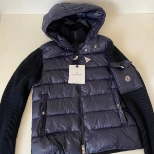 Mörkblå Moncler pufferjacka med huva - Snygg mörkblå pufferjacka från Moncler med stickade ärmar och vadderad kropp. Jackan har en stor huva, dragkedja framtill och Moncler-logga på ärmen. Perfekt för kyliga dagar och ger en riktigt clean look.