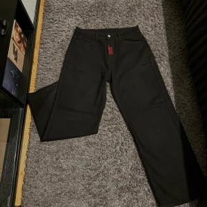 Svarta byxor med röd brodyr - Unika svarta byxor med röda broderade detaljer, inklusive text och motiv på bakfickorna samt stort mönster på ena benet. Raka ben och normal passform, tillverkade i bomull. Perfekta för dig som vill sticka ut med cool streetstyle.