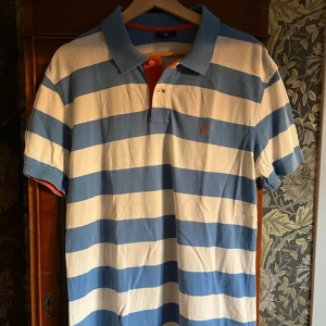 Randig pikétröja i blått och vitt - Snygg pikétröja från Gant med breda blå och vita ränder, klassisk krage och korta ärmar. Broderad logga på bröstet och orange detalj på insidan av knäppningen. Perfekt för en sportig och avslappnad stil.