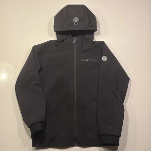 Svart vindjacka från Sail Racing - Svart vindjacka från Sail Racing med huva och hel dragkedja framtill. Jackan har diskret logga på bröstet och är tillverkad i ett slätt, vattenavvisande material. Muddar vid ärmslut och nederkant ger en snygg passform. Perfekt för blåsiga dagar.