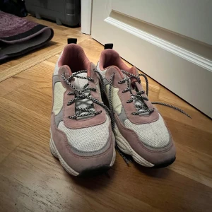 Vita och rosa sneakers - Snygga sneakers i vitt och ljusrosa. Skorna har mesh- och mockadetaljer, svartvitt mönstrade snören och en chunky sula. Perfekta för dig som gillar en sportig och trendig look. Bekväm passform och cool färgkombo.