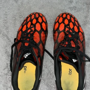 Adidas Predator fotbollsskor röd/svart - Säljer ett par Adidas Predator fotbollsskor i rött och svart med vita detaljer och mönstrad ovandel. Skorna har snörning och är tillverkade i syntetmaterial. Perfekta för dig som vill sticka ut på planen med grym design och bra grepp.
