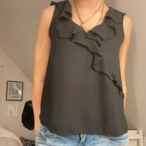 Svart ärmlös blus med volang - Svart ärmlös blus med snygg volangdetalj längs v-ringningen och framtill. Toppen har en lös passform och är tillverkad i ett lätt, flowy material som ger en skön känsla. Perfekt att styla med jeans eller kjol för en trendig look.