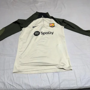 FC Barcelona långärmad träningströja Nike - Snygg långärmad träningströja från FC Barcelona med klubbmärke och Spotify-logga på bröstet. Tröjan är beige med mörkgröna ärmar och har halv dragkedja vid halsen. Tillverkad i lätt och funktionellt material, perfekt för fotbollsträning.