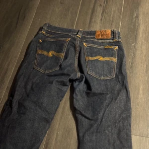 Mörkblå jeans från Nudie Jeans - Säljer ett par mörkblå jeans från Nudie Jeans med klassiska kontrastsömmar och snygga bakfickor med gul brodyr. Jeansen har fem fickor, bälteshällor och är tillverkade i slitstarkt denim. Perfekta för dig som gillar stilrena och tidlösa jeans. Lite dåliga bilder men kom privat för bättre och jeansen är lite så så dem är perfekt för dig som är lite mindre 