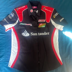 Ferrari Racing skjorta svart/röd - Racing-inspirerad skjorta från Ferrari med broderade loggor som Santander, Jinyu Tires och Ferrari Driver Academy. Skjortan är svart med röda och vita detaljer, korta ärmar och knappar framtill. Perfekt för motorsportfans som vill sticka ut med en sportig look.