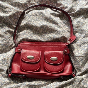 Röd axelväska med dubbla fickor - Snygg röd axelväska i skinn med två framfickor med silverfärgade knäppen och vit kontrastsöm. Väskan har justerbar axelrem och dragkedja upptill. Perfekt storlek för det viktigaste och ger en cool retrovibe till din outfit.