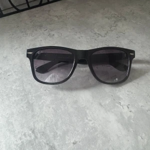 Svarta solglasögon Ray-Ban Wayfarer - Klassiska svarta Ray-Ban Wayfarer solglasögon med bred båge och mörka glas. Snygg och tidlös design med Ray-Ban-logga på sidan. Tillverkade i Italien och passar perfekt för dig som vill ha en ikonisk look.
