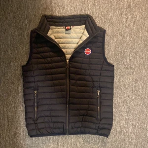 Dunväst från Colmar - Snygg svart dunväst från Colmar med quiltad design och hög krage. Västen har två fickor med dragkedja och klassisk Colmar-logga på bröstet. Perfekt för lager-på-lager och enkel att matcha med olika outfits. Litet litet hål precis vid en av dragkedjorna för fickorna. Se bild 4