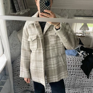 Beige rutig overshirt  - Säljer en oversized rutig overshirt från NA-KD i beige och grå toner. Jackan har stora bröstfickor med knappar, klassisk krage och är mjuk i materialet. Använd fåtal gånger.