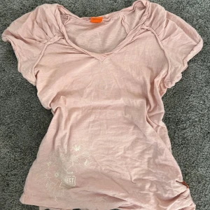 Vintage t-shirt  - Ljusrosa vintage -shirt med v-ringad hals och korta ärmar. T-shirten har diskret blommönster framtill och små knappar på sidan. 