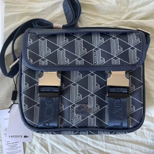 Lacoste Väska  - Snygg helt oanvänd blå axelväska från Lacoste med geometriskt mönster i vitt. Väskan har justerbar axelrem med Lacoste-logga, två metallspännen framtill och praktiskt lock. Materialet är slitstark polyester och designen är modern och sportig.