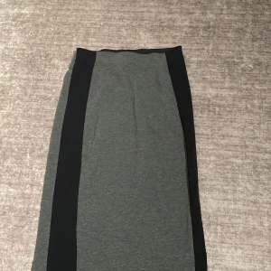 Grå och svart långkjol från Gina Tricot - Snygg långkjol från Gina Tricot i storlek S. Kjolen är grå med svarta paneler längs sidorna och har en rak, enkel design. Perfekt för dig som gillar stilrena plagg med en modern touch.