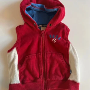 Röd väst med huva från H&M L.O.G.G. - Sportig röd väst med huva från H&M L.O.G.G. för baby, med vita paneler på sidorna, blått innerfoder i huvan och tryck med texten 'BGSA' och en fotboll på bröstet samt 'Football' på ryggen. Dragkedja framtill och två fickor.