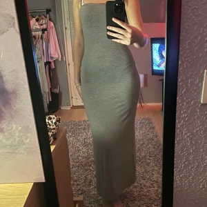 Grå långklänning med smala axelband - Säljer en stilren grå långklänning med smala axelband och rak passform. Klänningen är figurnära och har en enkel, minimalistisk look som passar perfekt för dig som gillar en clean stil. Materialet känns mjukt och stretchigt för en bekväm känsla.