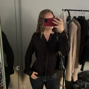 Svart prickig blus från Noa Noa - Svart blus från Noa Noa med diskreta prickar i vitt och rött. Långärmad modell med lätt rynkade detaljer och v-ringning. Knappar hela vägen. Mer som en M