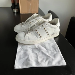 Valentino Rockstud vita sneakers - Säljer ett par vita Valentino Rockstud sneakers i storlek 42 men fin 41 i skinn med ikoniska nitar i silver längs sidorna men en nita är borta som man kan se på bild 5. Klassisk rund tå, snörning och platt sula. Kommer med original dustbag. Utan box😊
