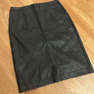Skinn kjol från KappAhl - Snygg svart skinn kjol från KappAhl. Kjolen har en markerad midja, dragkedja med ringdetalj bak och en liten slits för extra rörelsefrihet. Perfekt för dig som gillar en stilren och edgy look. Den kanske behöver strykas annars har den inga defekter🖤