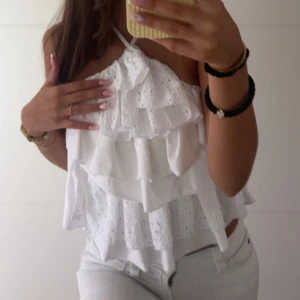 Vit volangig ärmlös linnetopp - Superfin vit linnetopp med flera lager volanger och broderade detaljer. Toppen är ärmlös med smala axelband som knyts i nacken för en halterneck-look. Perfekt för en fräsch och somrig stil.