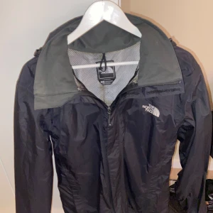 The North Face svart vindjacka - Säljer en svart vindjacka från The North Face med huva och meshfoder. Jackan har dragkedja framtill, hög krage och logga på bröstet. Perfekt för blåsiga dagar och har en clean, sportig look. Passar dig som gillar outdoor och streetstyle.