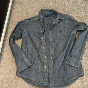 Blå skjorta från Ralph Lauren - Snygg blå skjorta från Ralph Lauren med klassisk krage och knappar framtill. Skjortan har lång ärm och ikoniska broderade loggan på bröstet. Tillverkad i mjuk bomull och passar perfekt till jeans eller chinos.