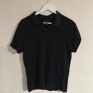 Svart pikétröja - Klassisk svart pikétröja från H&M i ribbad bomull. Tröjan har korta ärmar, krage och knappar framtill. Perfekt basic-plagg med stilren look och bekväm passform. Storleken är M men funkar även till S. Skriv vid minsta fundering🍾🌠