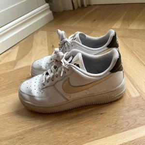 Nike Air Force 1 vita sneakers - Säljer ett par klassiska Nike Air Force 1 sneakers i vitt med beiga detaljer och brun häl. Skorna har snörning, perforerad tå och platt sula. Perfekta för dig som gillar en clean och tidlös look med en twist. Materialet är skinn och syntet.