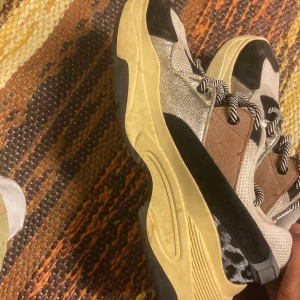 Chunky sneakers i beige, svart och guld - Säljer ett par chunky sneakers med coola detaljer i beige, svart och guld. Skorna har grov sula, snörning med svartvita skosnören och inslag av mesh, mocka och metallic. Perfekta för dig som gillar statement-skor med unik look.