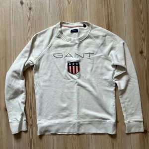 Vit sweatshirt från GANT - S - Vit sweatshirt från GANT med broderad logga och sköld på bröstet. Klassisk rund halsringning och långa ärmar. Tröjan är i mjuk bomull och har ribbade muddar vid ärmslut och nederkant. Perfekt för en clean och stilren look.