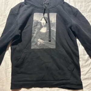 Svart hoodie Mona Lisa The Cool Elephant - Svart hoodie från The Cool Elephant med ikoniskt Mona Lisa-tryck på bröstet. Tröjan har huva med snörning och en stor magficka. Perfekt för dig som gillar streetstyle och konstnärliga detaljer. 12-14 år