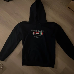 Svart hoodie Monte Carlo Kartisto - Svart hoodie från Kartisto med Monte Carlo-tryck och färgglada flaggor på bröstet. Klassisk huva med snörning och långa ärmar. Perfekt för dig som gillar en stilren och sportig look.
