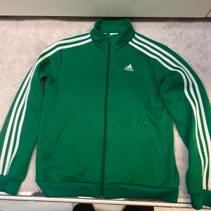 Grön Adidas ziptröja med vita ränder - Grön Adidas ziptröja med klassiska vita ränder längs ärmarna och broderad logga på bröstet. Hög krage, dragkedja hela vägen och två fickor med dragkedja. Perfekt för en sportig look eller chill vardagsstil.