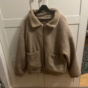 Beige teddyjacka från Lily Lulu - Mysig beige teddyjacka från Lily Lulu med dragkedja framtill och två stora fickor. Jackan har en klassisk krage och är tillverkad i fluffigt teddy-material som håller dig varm under kyliga dagar. Perfekt för dig som gillar en chill och trendig look. Den är väldigt stor i storleken.