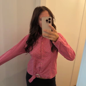 Rosa randig skjorta med knytband - Rosa skjorta med tunna vita ränder och snyggt knytband nertill. Den är inte strykt 😬 Klassisk krage, knäppning framtill och lång ärm. Skjortan har en lite loose passform och är gjord i mjuk bomull. AS SNYGG men passar inte riktigt resten av min garderob så aldrig använt. Köp för 500kr